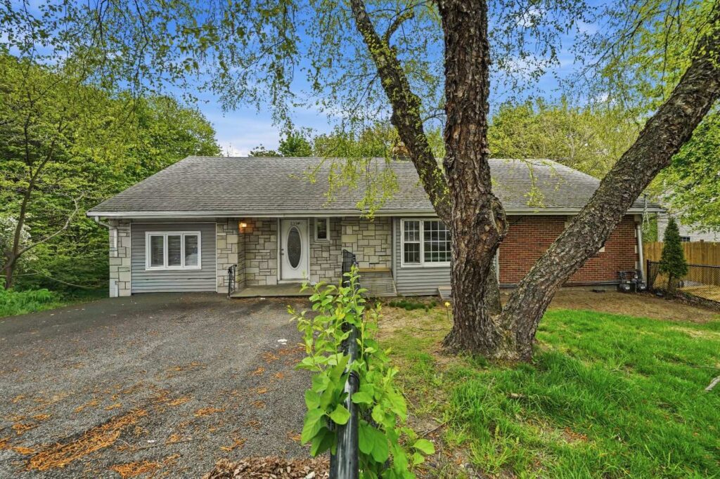 Front view of a single-story home with mixed stone, siding, and brick exterior, surrounded by mature trees, a gravel driveway, and a grassy front yard.